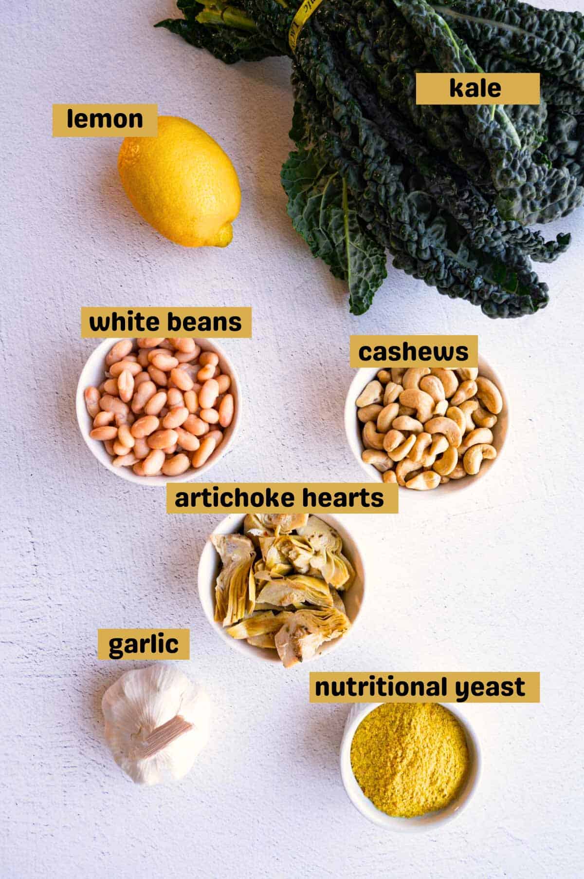 White beans, nutritional yeast, garlic, artichoke hearts, cashews, white beans, lemon, and kale on a gray backdrop.