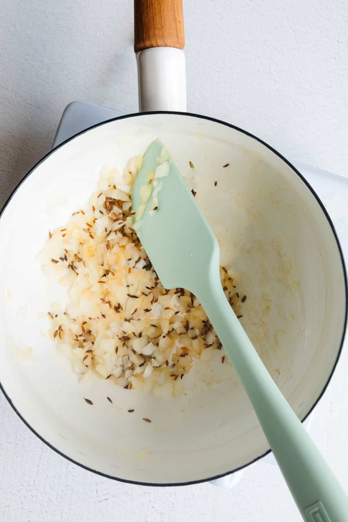 Onion and cumin seeds cooking in a white saucepan.