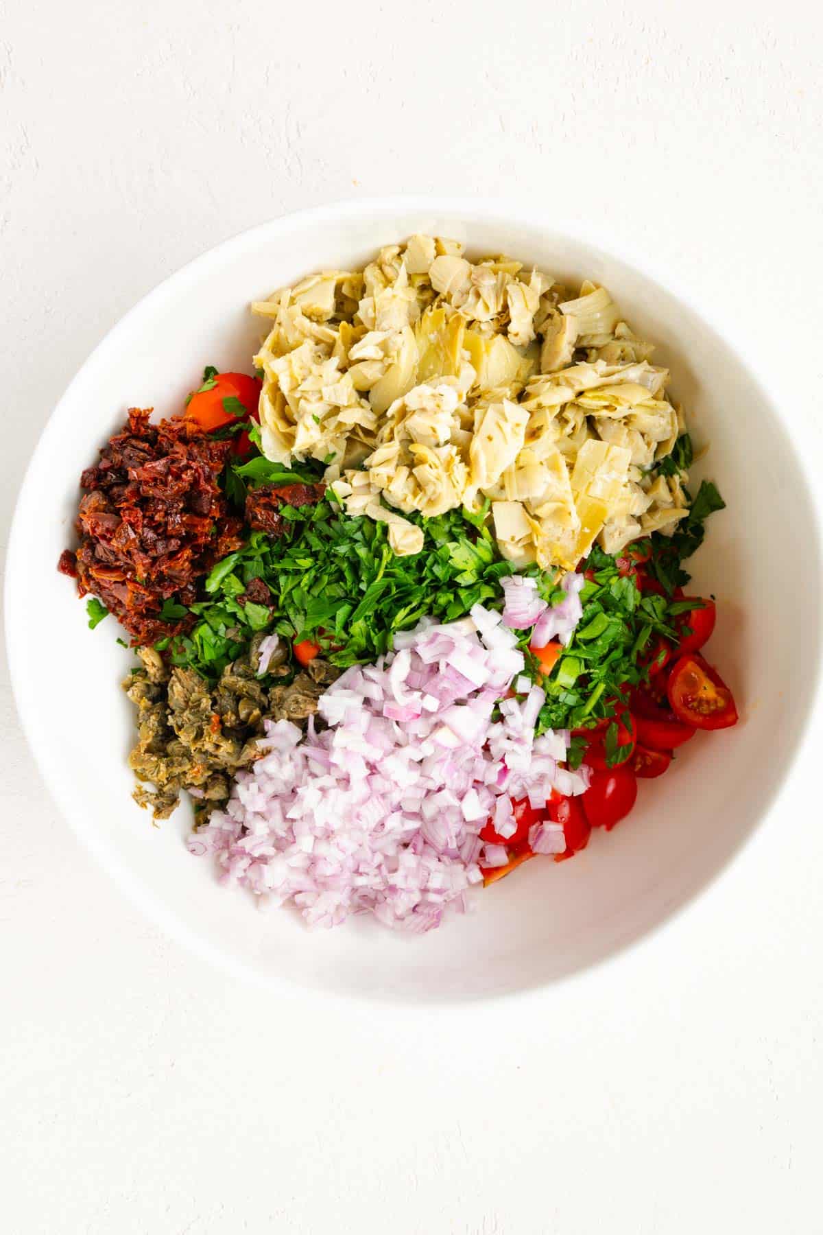 Quartered cherry tomatoes, chopped sun-dried tomato, shallots, capers, parsley, and artichoke hearts in a white bowl.