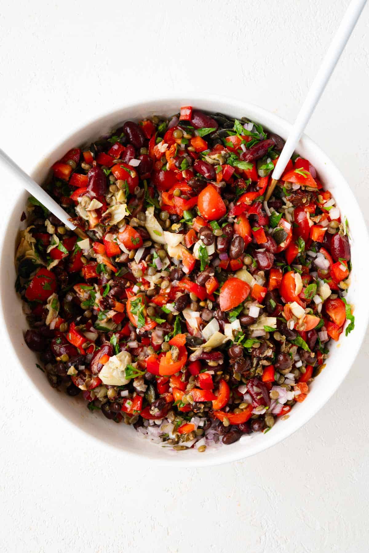 Dense bean salad with kidney beans, black beans, lentils, tomatoes, parsley, shallots, artichoke hearts, and capers mixed and chopped in a white bowl.