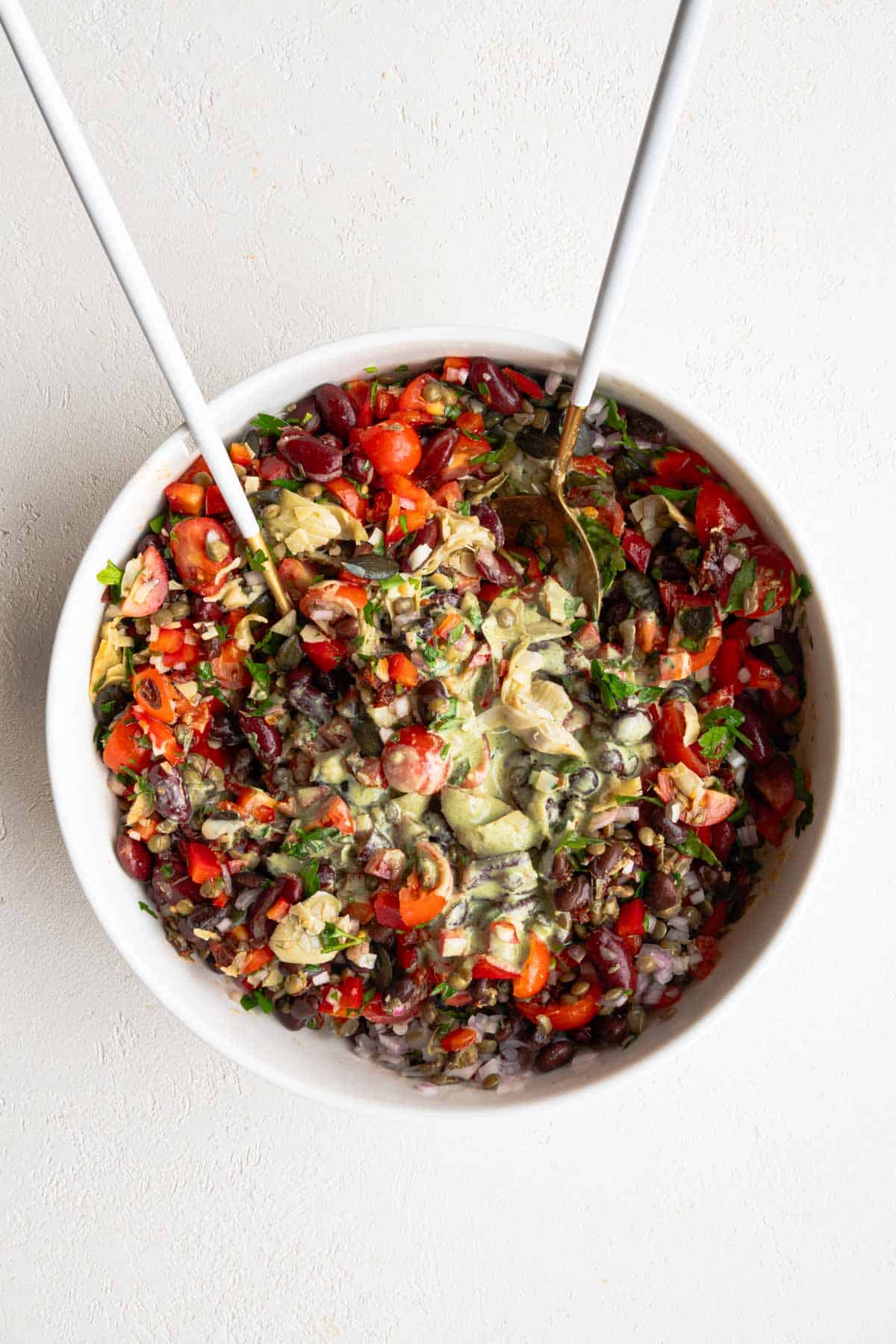 Kidney beans, black beans, lentils, tomatoes, parsley, shallots, artichoke hearts, and capers with green dressing in a white bowl.