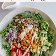 Arugula, quinoa, chickpeas, artichoke hearts, capers, tomatoes, cucumber and hummus dressing in a white bowl.