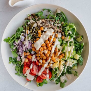 Arugula, quinoa, chickpeas, artichoke hearts, capers, tomatoes, cucumber and hummus dressing in a white bowl.