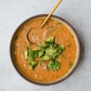 Vegan barley soup with white beans and vegetables in a bowl with a spoon topped with avocado.