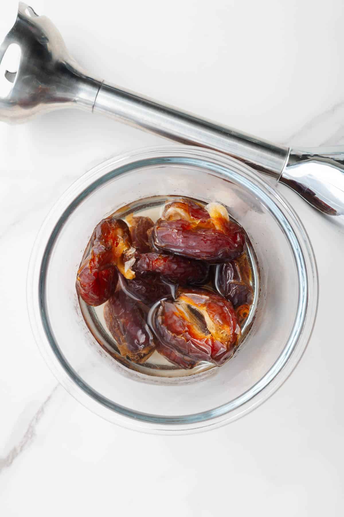 Medjool dates in water in a glass jar next to an immersion blender.