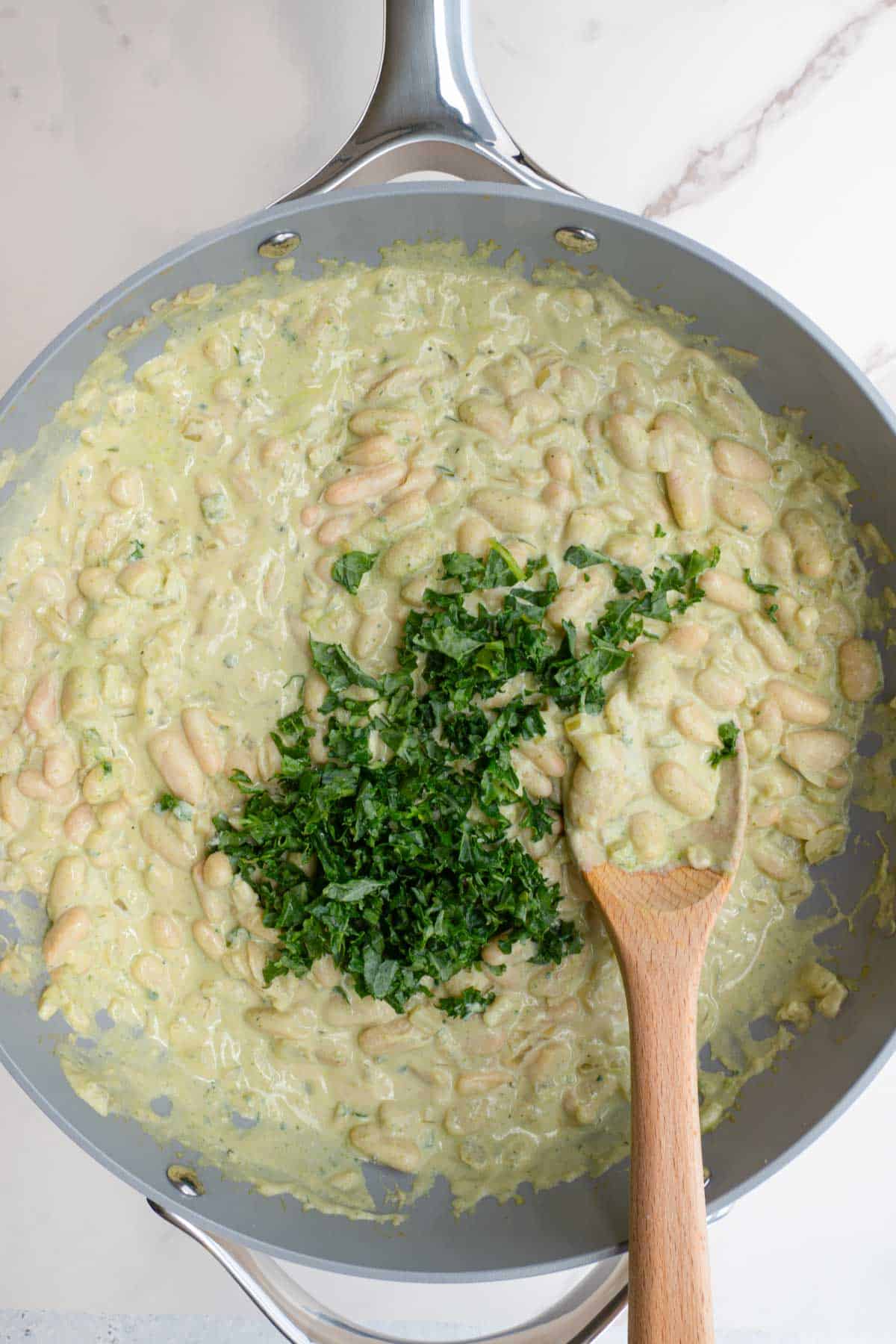 Creamy white beans with greens in a grey saucepan with a wooden spoon.