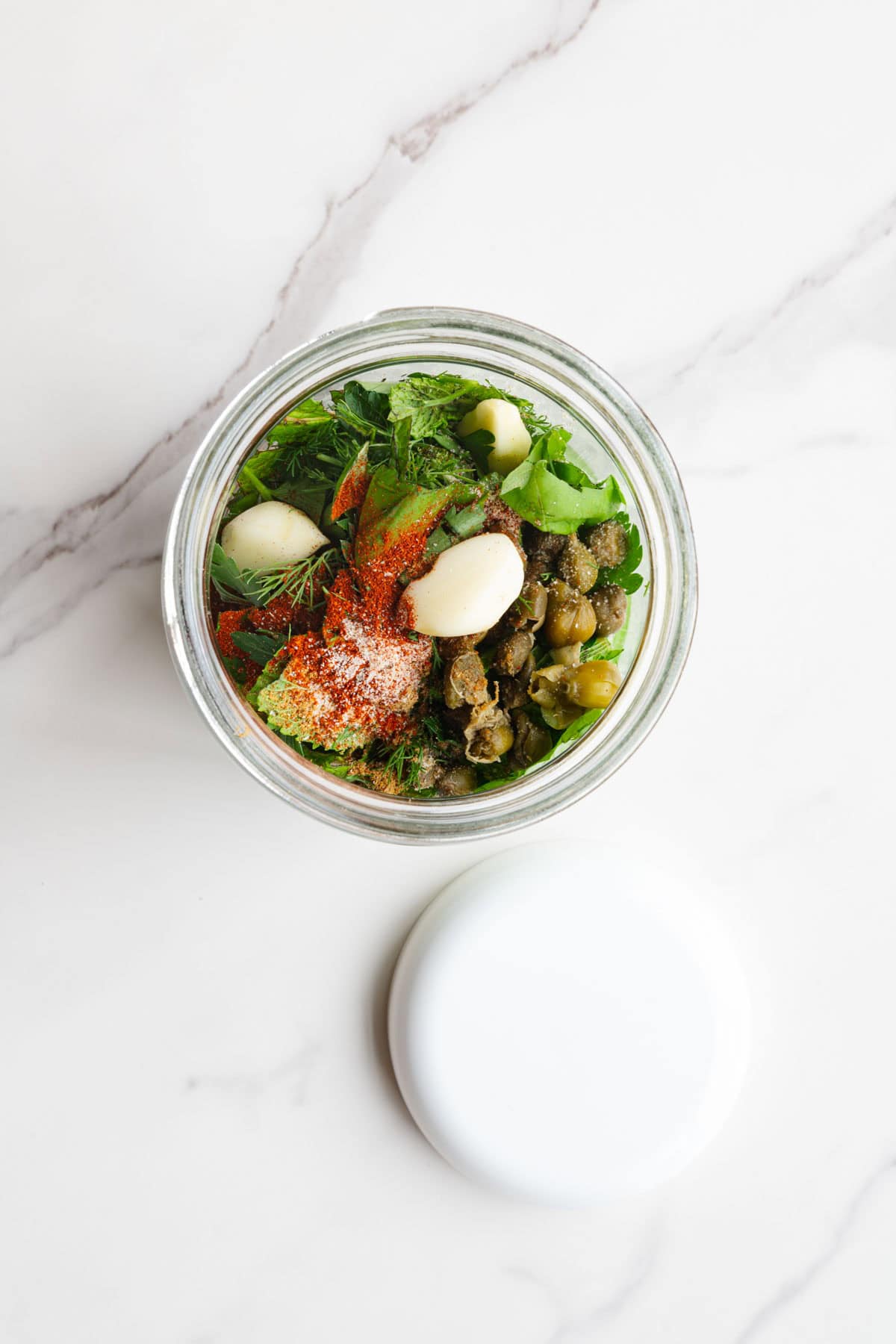 Capers, garlic, fresh herbs, and seasoning in a glass jar with a white lid.