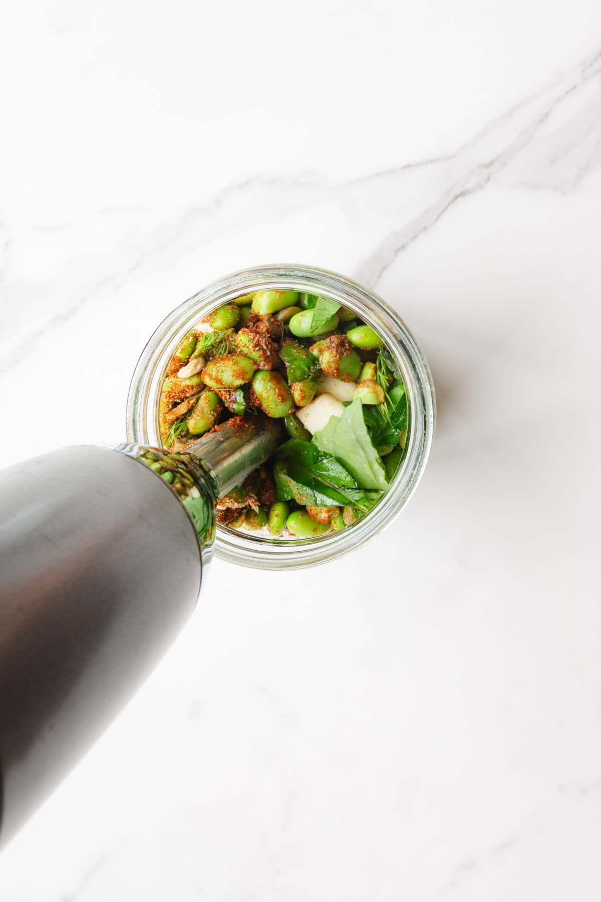 Edamame, green peas, and seasonings being blended with an immersion blender in a glass jar.