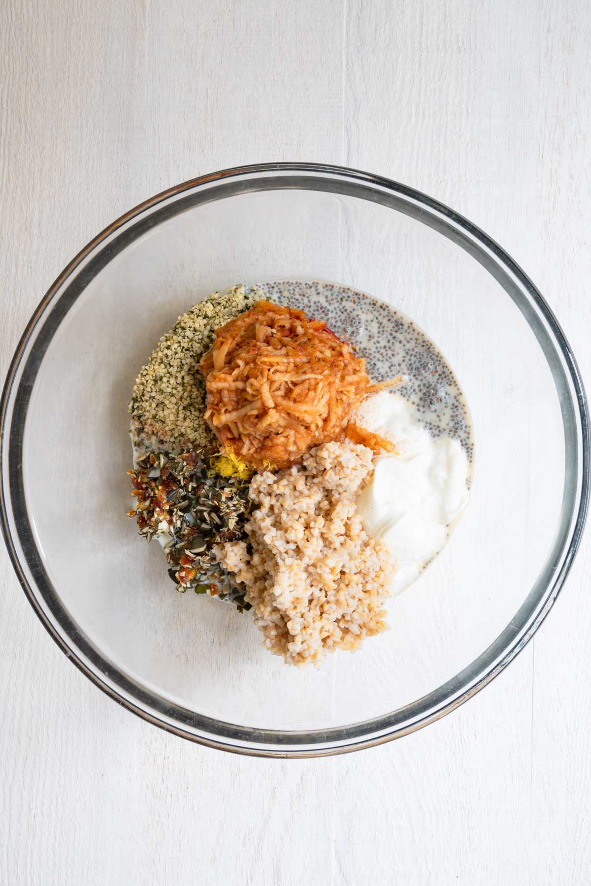 Chia seeds, steel-cut oats, hemp hearts, dates, vanilla, nondairy milk, pumpkin seeds, cinnamon, yogurt, lemon, and grated apple in a glass bowl.
