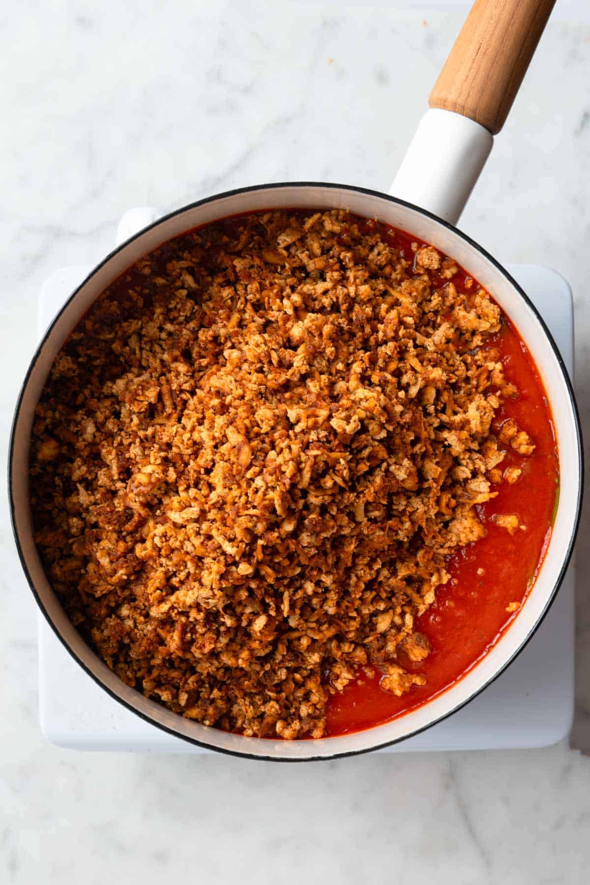 Crumbled tofu on simmering marinara sauce in a white saucepan.