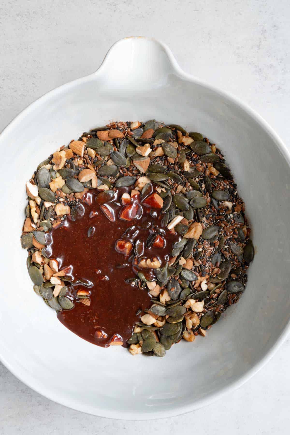 Nuts and seeds with warm date syrup and tahini for grain-free granola in a large bowl.