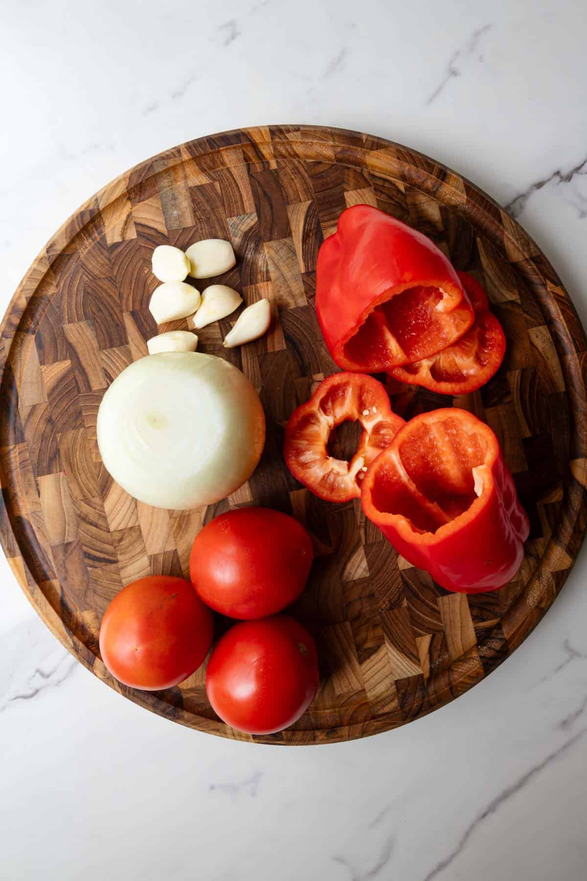 Tomatoes, red bell pepper, onion, and garlic on a wooden cutting board.