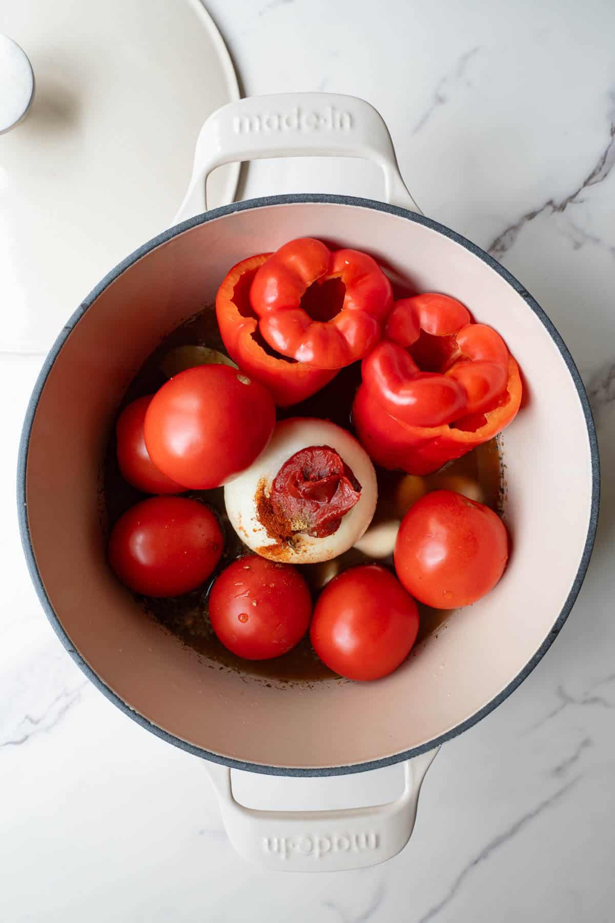 Tomatoes, red bell pepper, onion, tomato paste, seasoning, and garlic in a white saucepan.