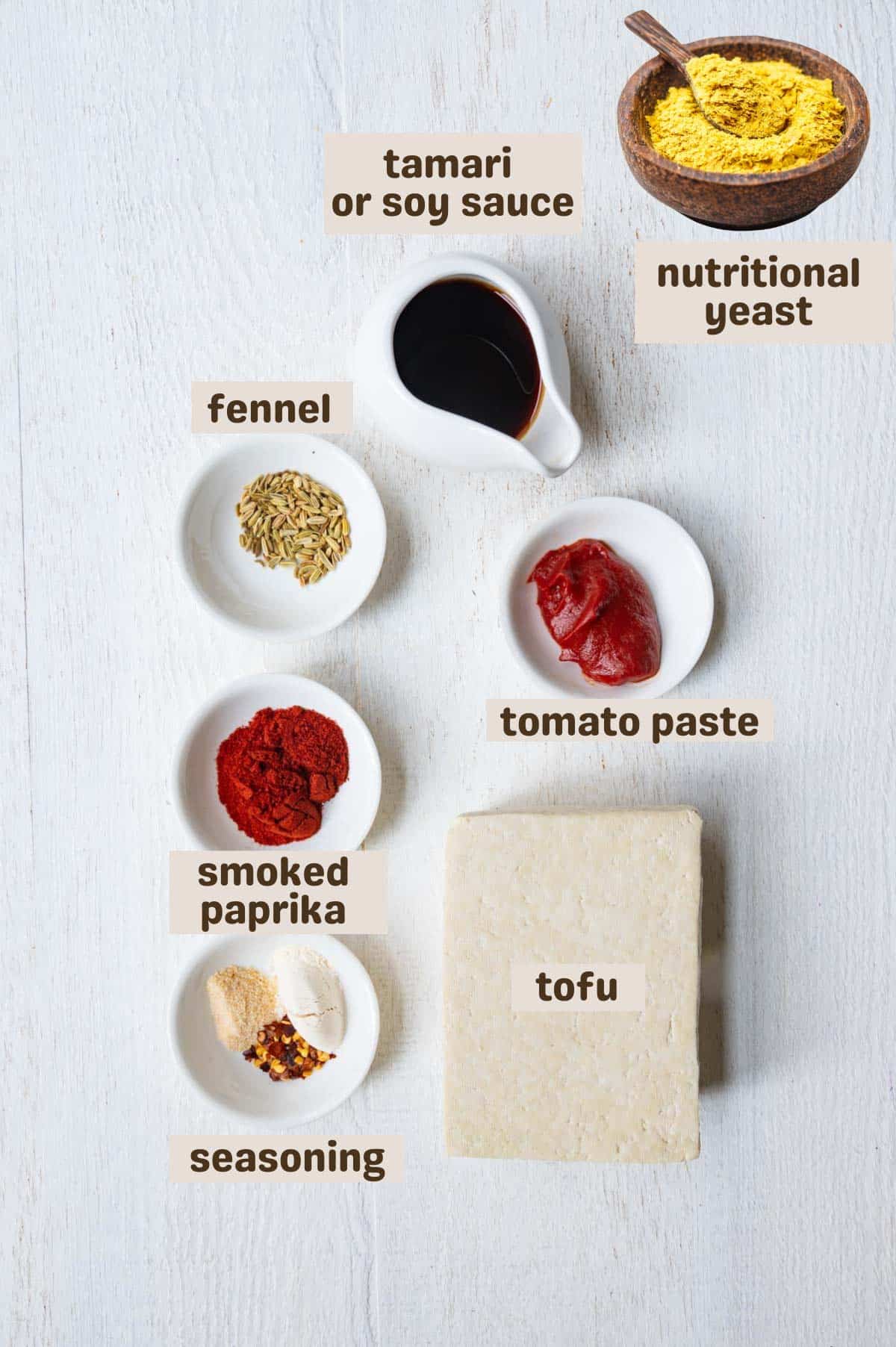 Chunks of tofu on a white baking sheet with smoked paprika, tomato paste, garlic powder, onion powder, crushed red pepper, nutritional yeast, and whole fennel seeds.