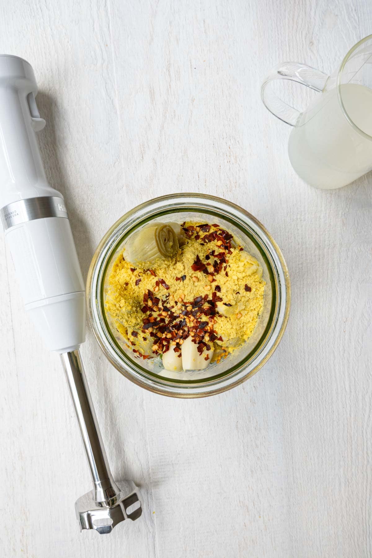 Nutritional yeast, miso, artichoke hearts, date syrup, garlic, red pepper flakes, spinach, and lemon juice in a large glass jar next to an immersion blender and pasta water.