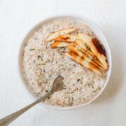 High-protein apple cinnamon chia seed oatmeal in a bowl with a spoon and sliced apples on top with a drizzle of date syrup.