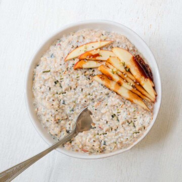 High-protein apple cinnamon chia seed oatmeal in a bowl with a spoon and sliced apples on top with a drizzle of date syrup.