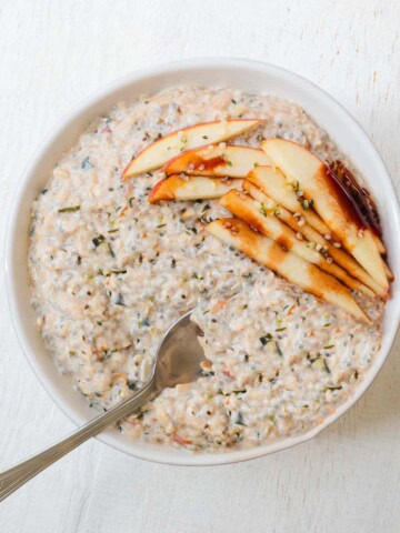 High-protein apple cinnamon chia seed oatmeal in a bowl with a spoon and sliced apples on top with a drizzle of date syrup.