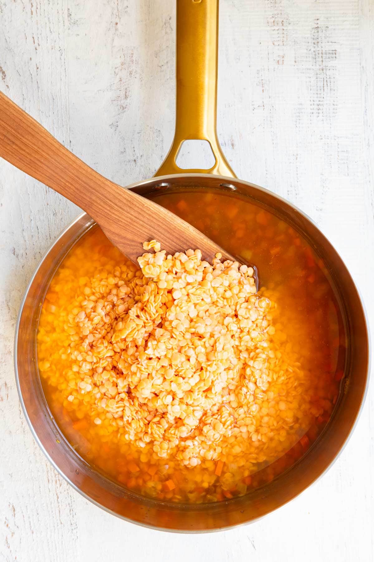 Cooked chopped onion and carrots, broth, and red lentils in a stainless saucepan.