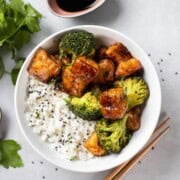 Tofu and broccoli teriyaki in a white bowl with rice and chopsticks.