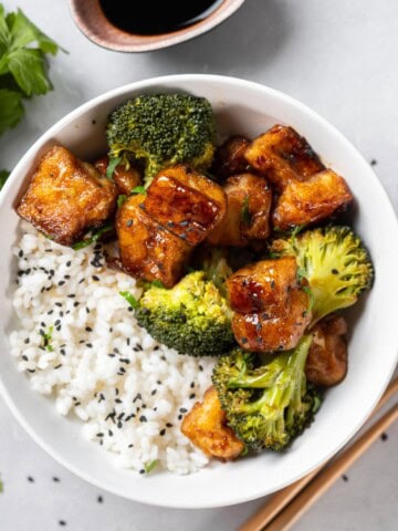 Tofu and broccoli teriyaki in a white bowl with rice and chopsticks.