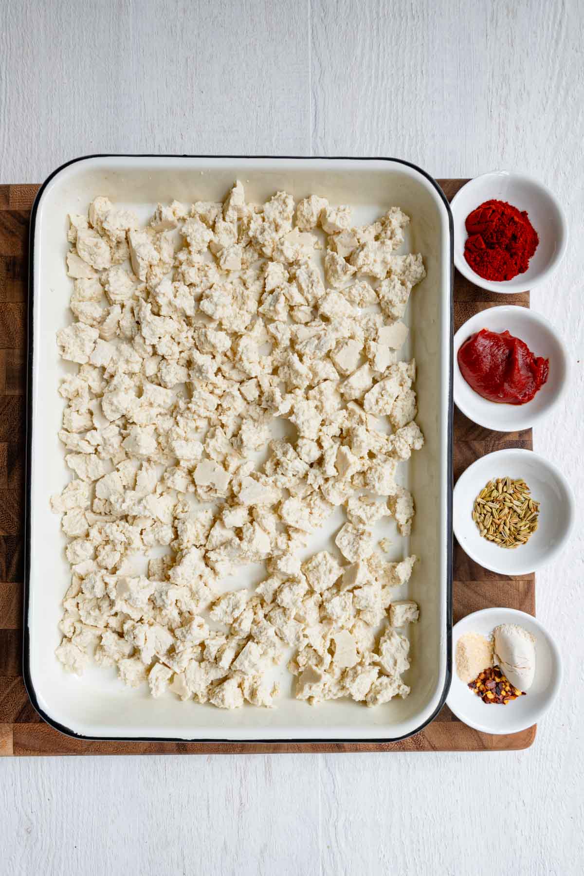 Chunks of tofu on a white baking sheet with smoked paprika, tomato paste, garlic powder, onion powder, crushed red pepper, and whole fennel seeds.