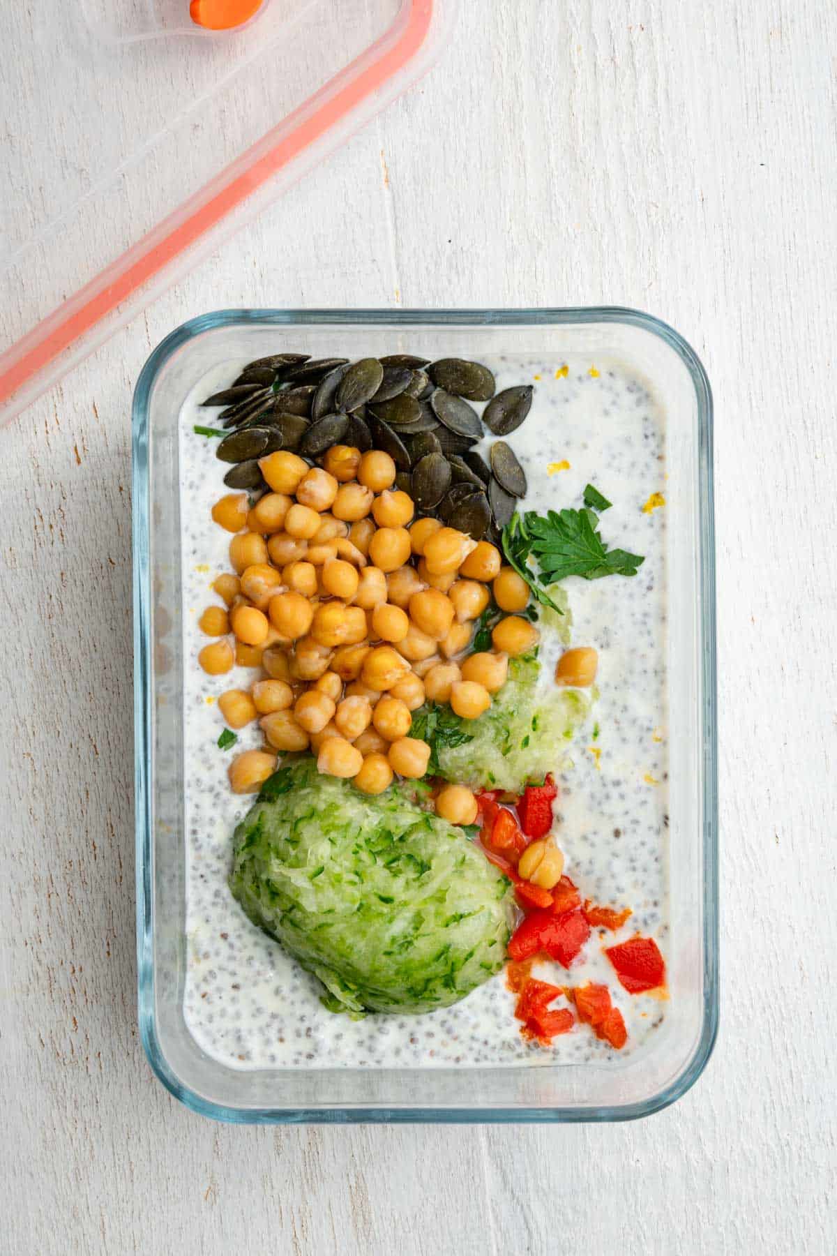 Chickpeas, pumpkin seeds, chopped roasted red pepper, grated cucumber, chopped parsley, and garlic on creamy chia pudding with yogurt in a glass container.