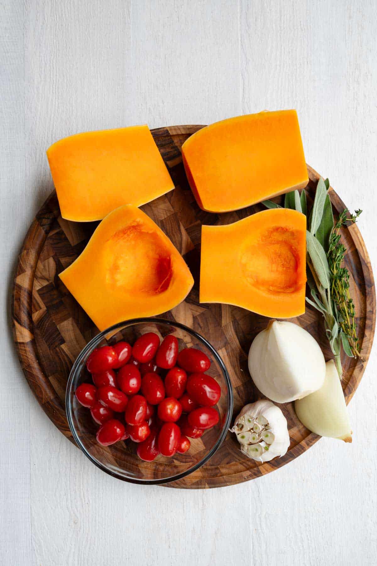 Prepped butternut squash, onion, cherry tomatoes, garlic, and fresh sage and thyme on a wooden cutting board.