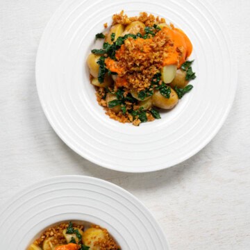 Two loaded potato bowls with sliced baby potatoes, tofu crumbles, chopped kale, and red sauce in white bowls.