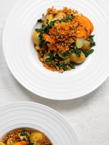Two loaded potato bowls with sliced baby potatoes, tofu crumbles, chopped kale, and red sauce in white bowls.
