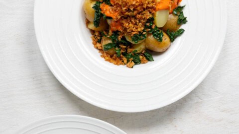 Two loaded potato bowls with sliced baby potatoes, tofu crumbles, chopped kale, and red sauce in white bowls.