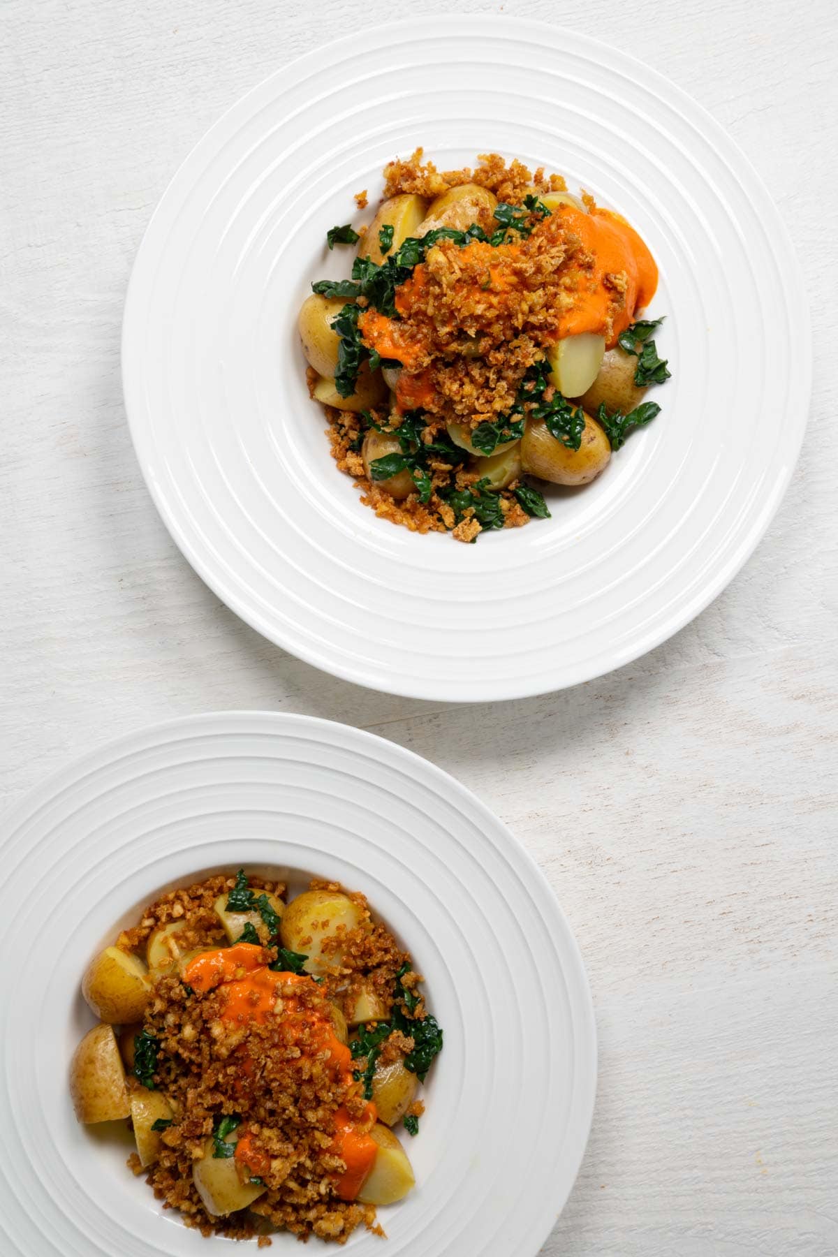 Two loaded potato bowls with sliced baby potatoes, tofu crumbles, chopped kale, and red sauce in white bowls.