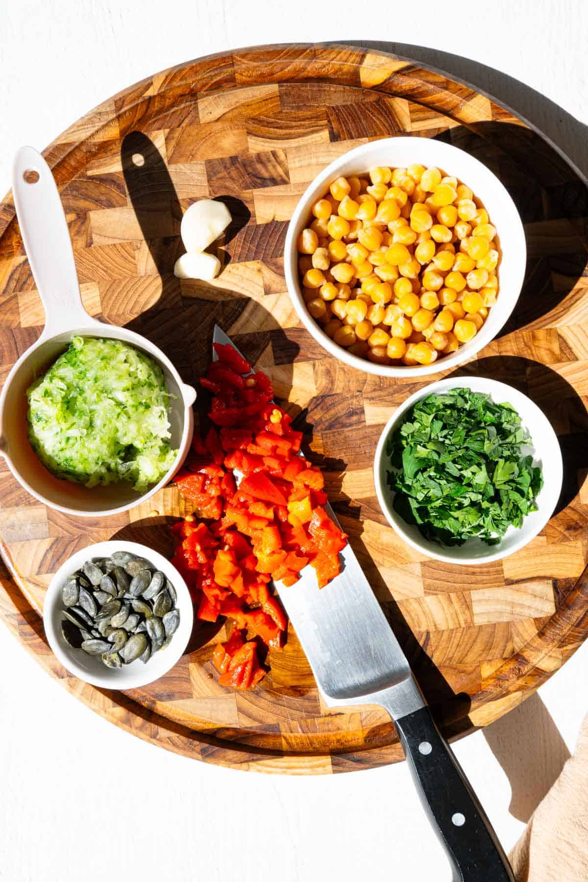 Chickpeas, pumpkin seeds, chopped roasted red pepper, grated cucumber, chopped parsley, and garlic on a wooden cutting board.