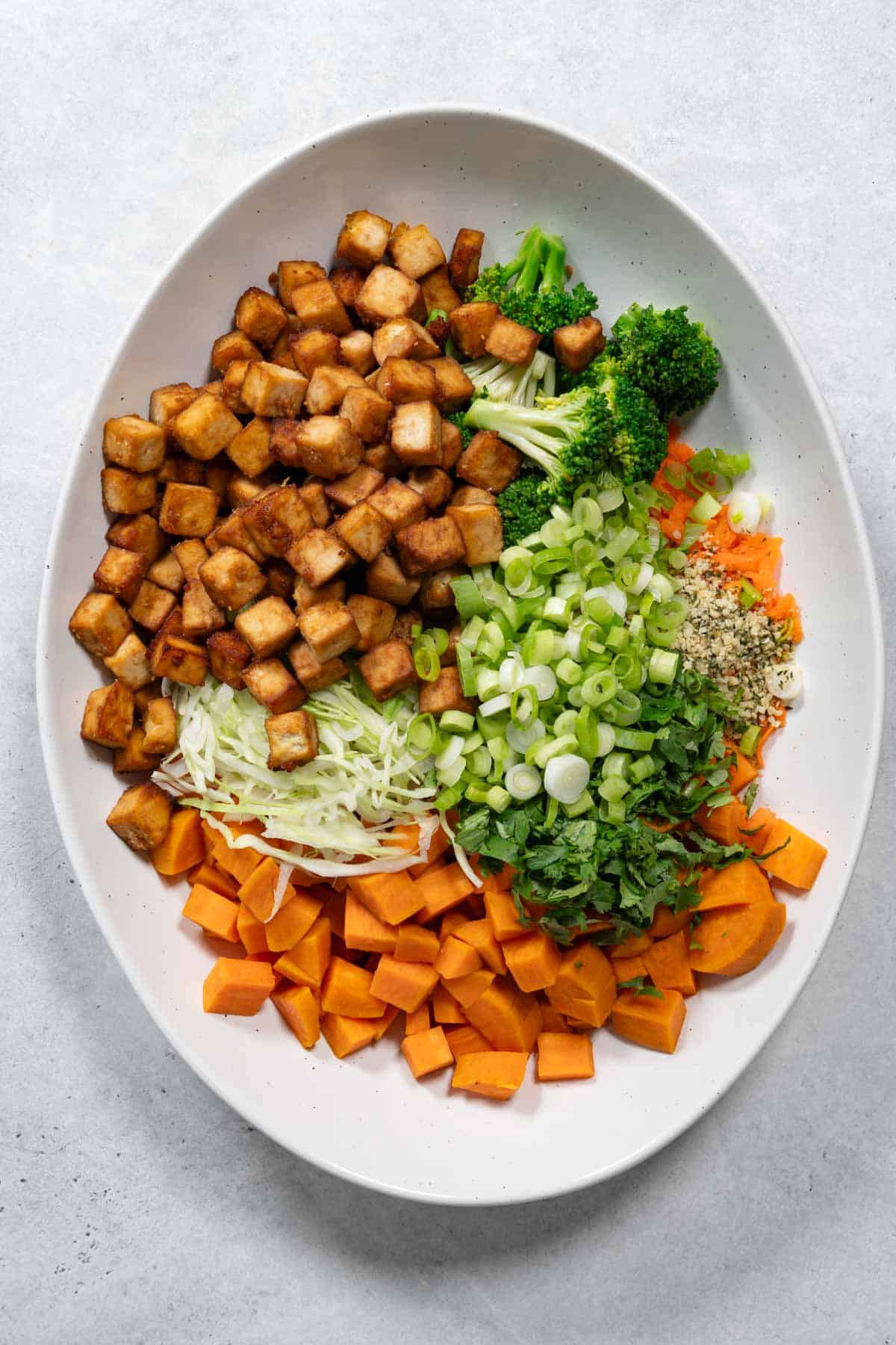 Cubed sweet potato, grated carrots, broccoli, sliced cabbage, hemp hearts, cilantro, tofu cubes, and green onion on a white plate.