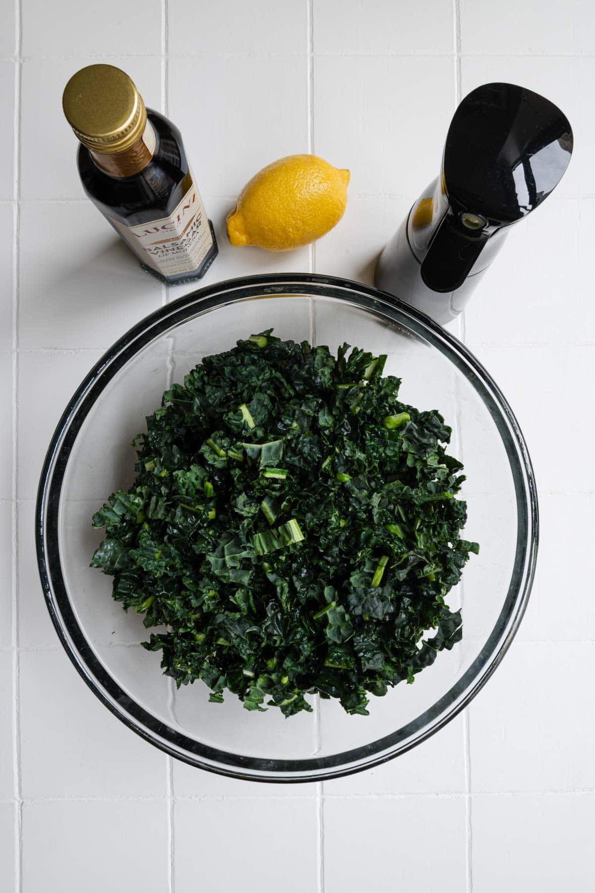Chopped kale in a large bowl with olive oil spray bottle, lemon, and balsamic vinegar.