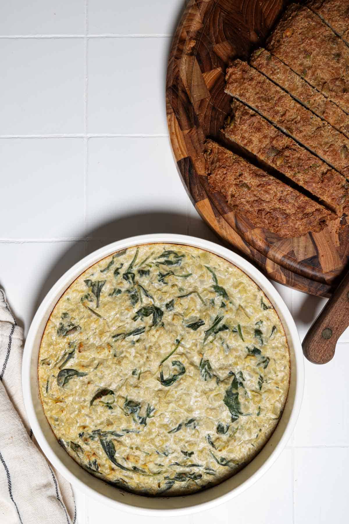 Creamy vegan spinach and artichoke dip in a round baking dish with sliced quinoa bread.