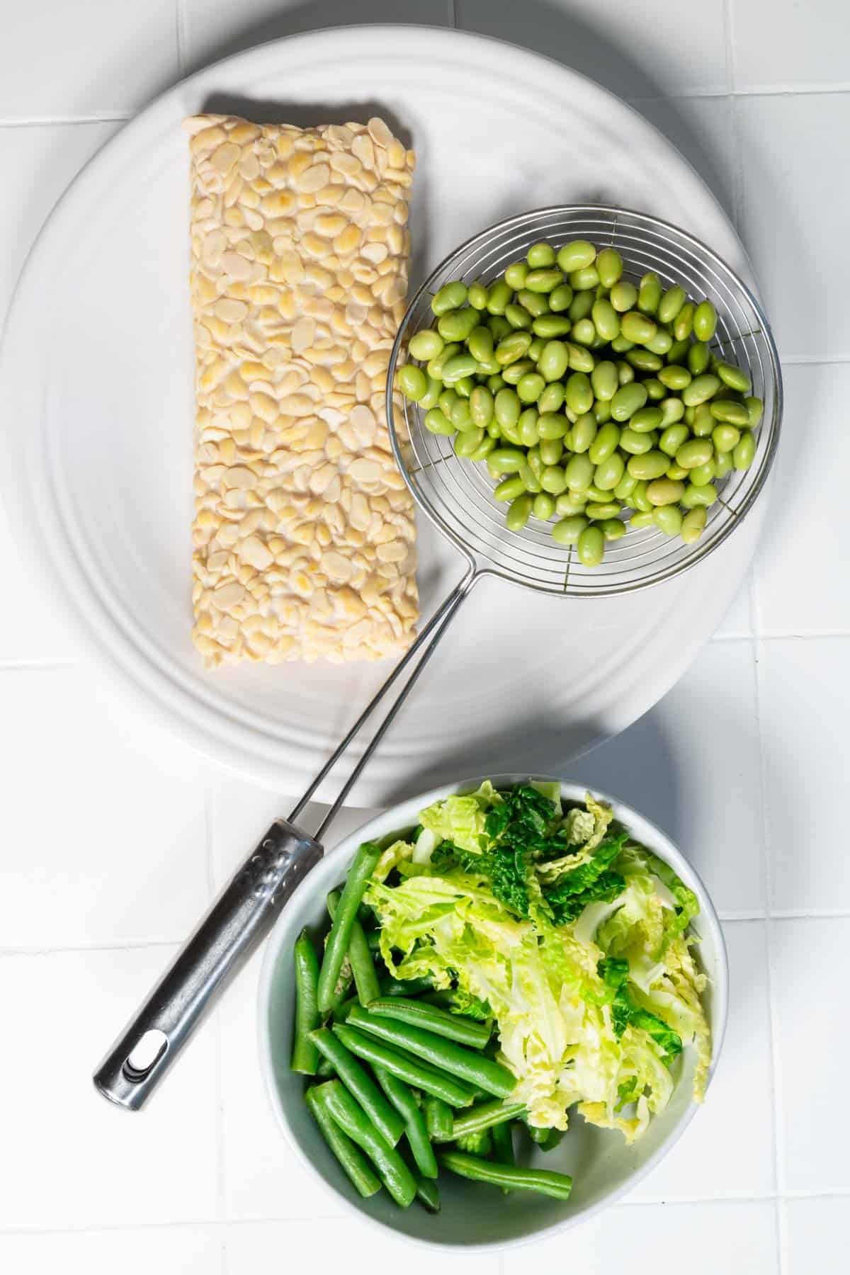 Simmered tempeh and edamame on a white plate and a bowl of steamed cabbage and green beans.
