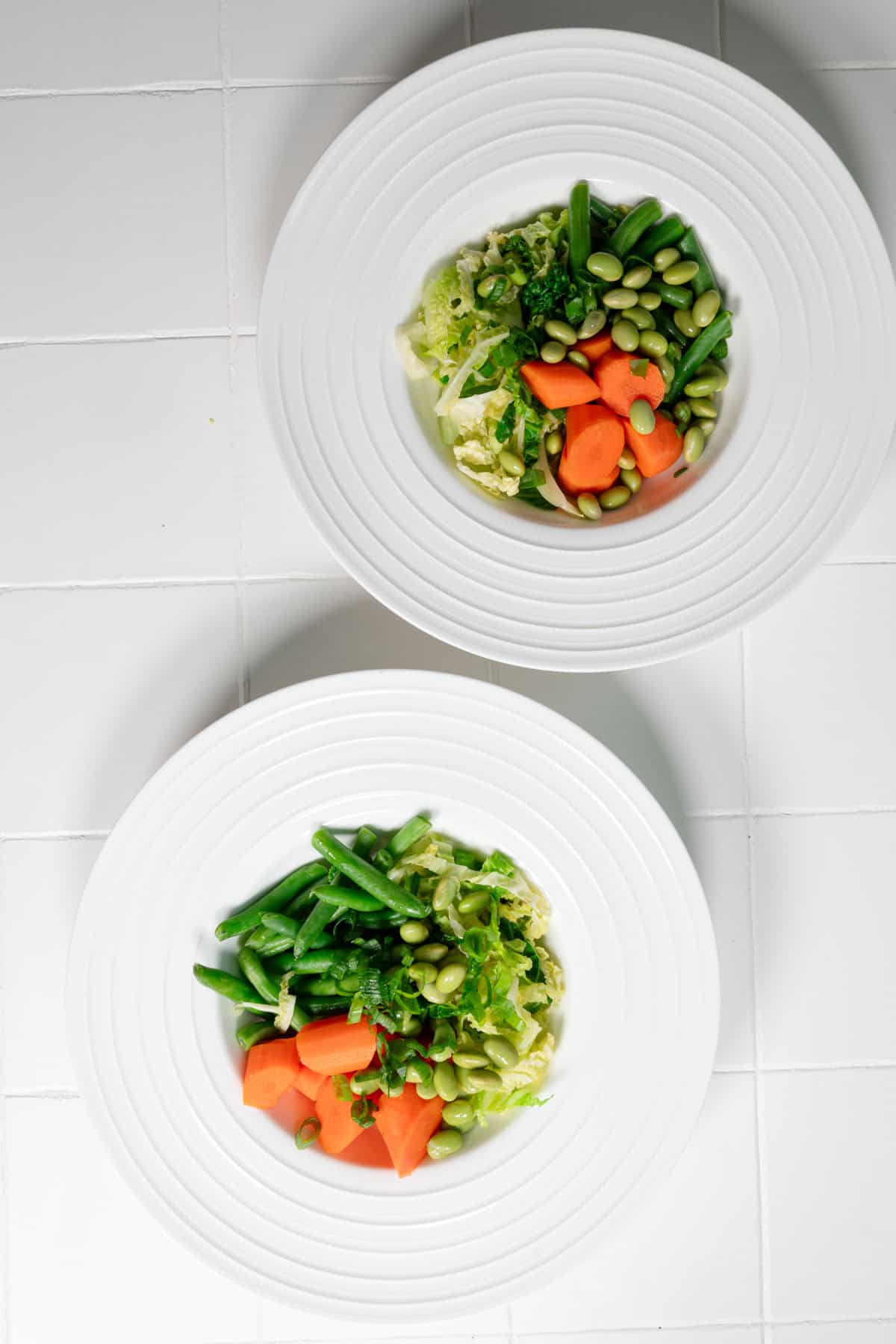 Steamed carrots, cabbage, green beans, and edamame in two white bowl.
