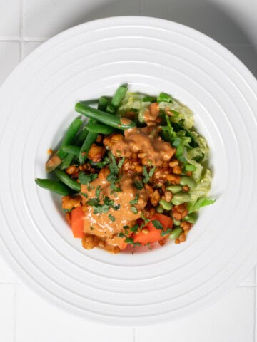 Steamed vegetable bowl with carrots, cabbage, edamame, tempeh, green beans, and peanut sauce in a white bowl.