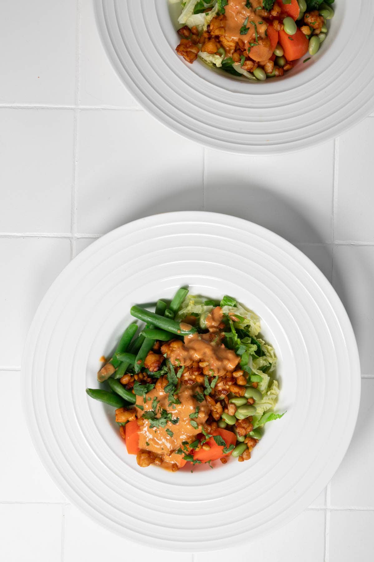 Steamed vegetable bowl with carrots, cabbage, edamame, tempeh, green beans, and peanut sauce in two white bowls.