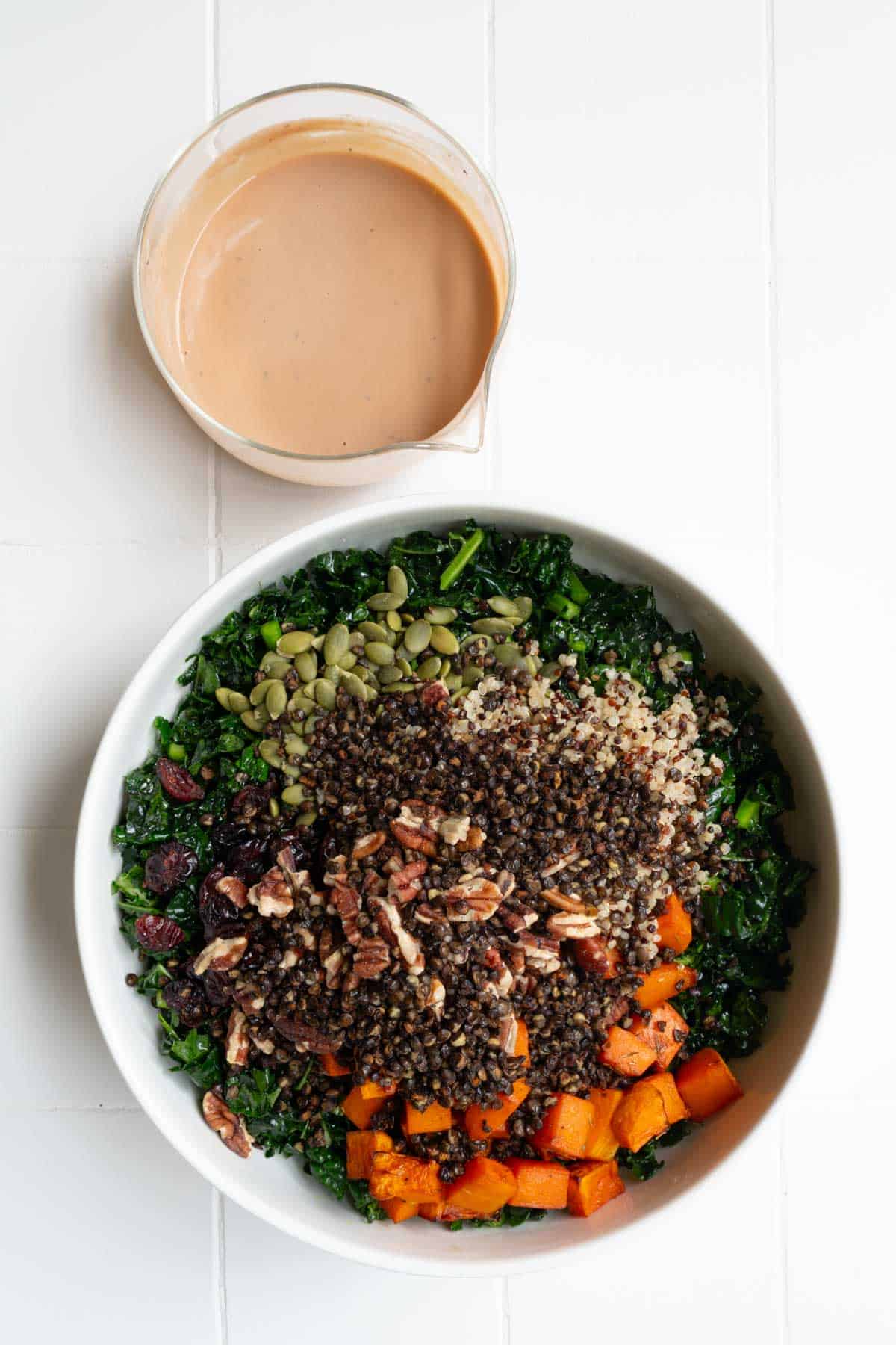 Kale, butternut squash, pecans, dried cranberries, quinoa, pepitas, and roasted lentils in a white bowl with creamy tahini dressing.