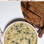 Creamy vegan spinach and artichoke dip in a round baking dish with sliced quinoa bread.