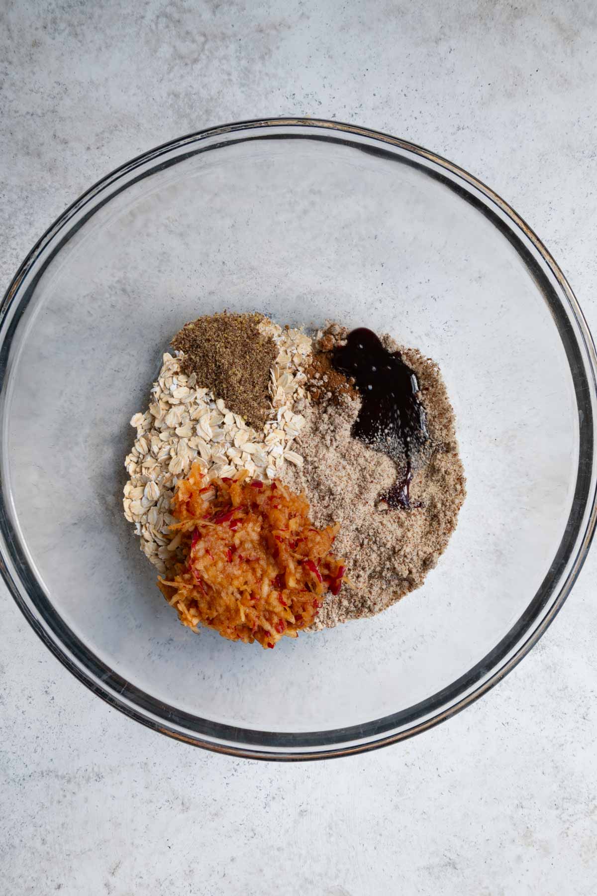 Rolled oats, grated apple, date syrup, flaxseed meal, cinnamon, and almond flour in a large glass bowl.
