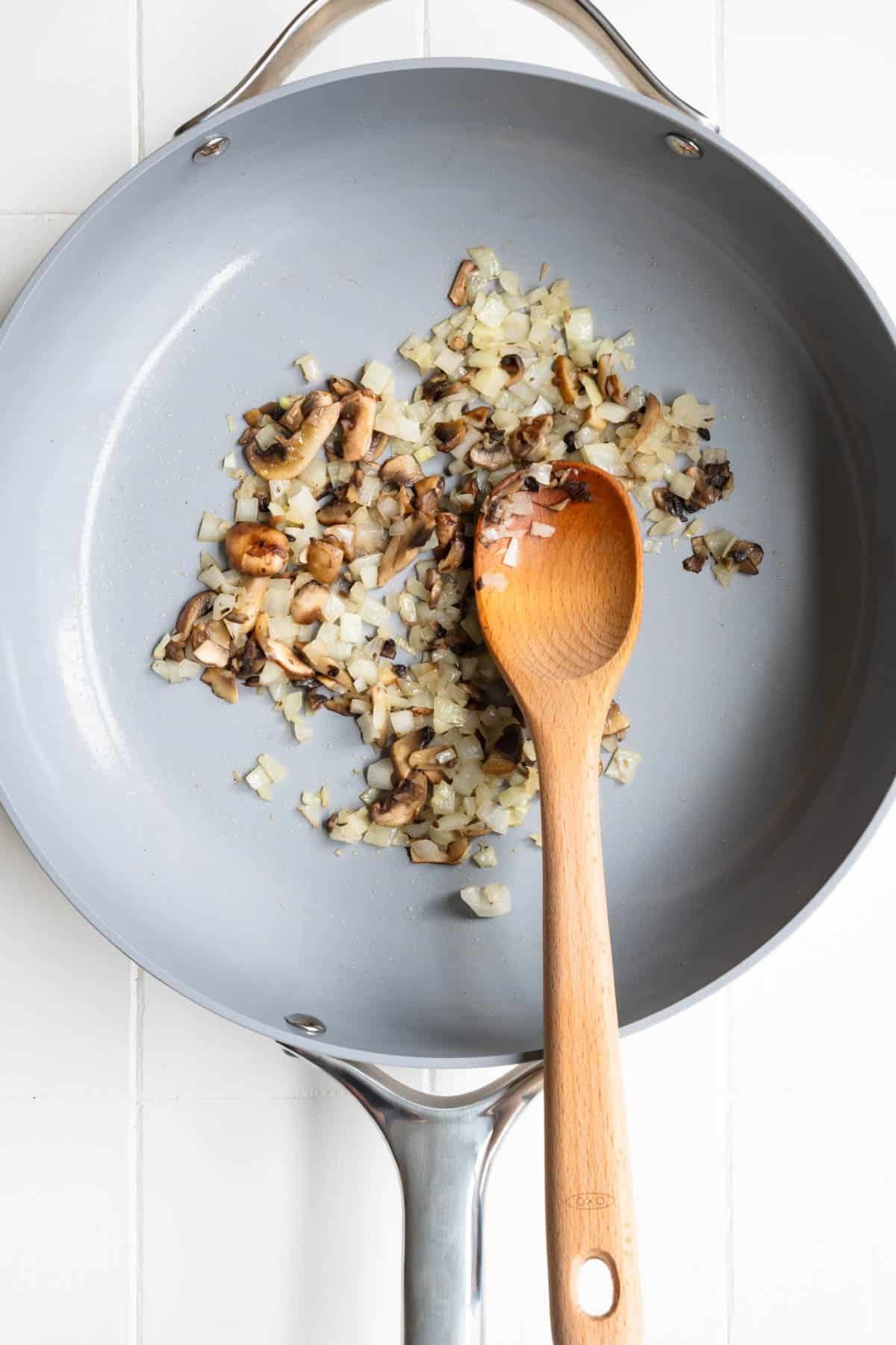 Cooked onion and mushrooms in a skillet with a wooden spoon.