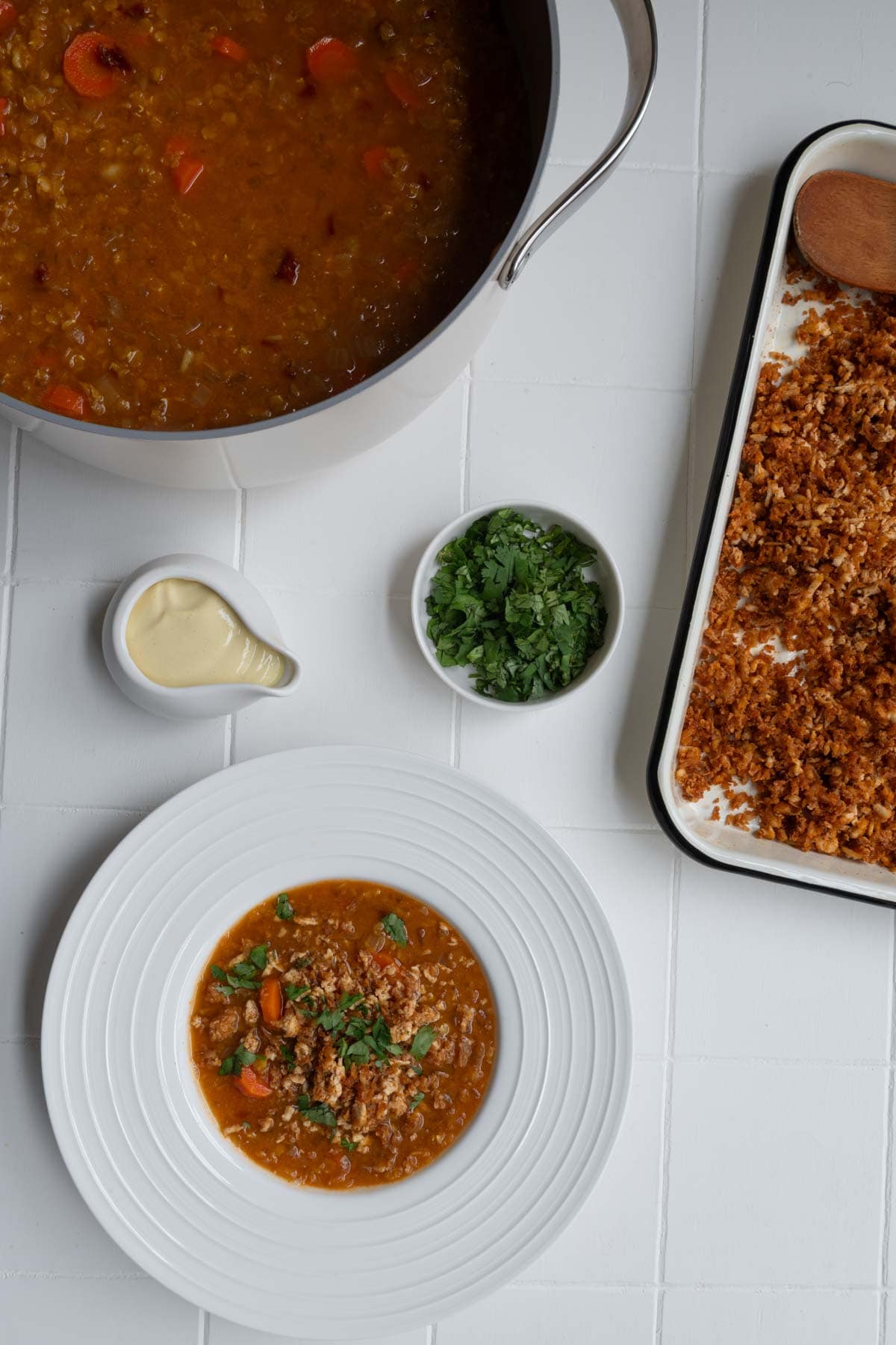 Tomato lentil soup with tofu cream and crumbled tofu in a white bowl with a baking sheet of crumbled tofu, chopped cilantro, a pot of soup, and tofu cream.
