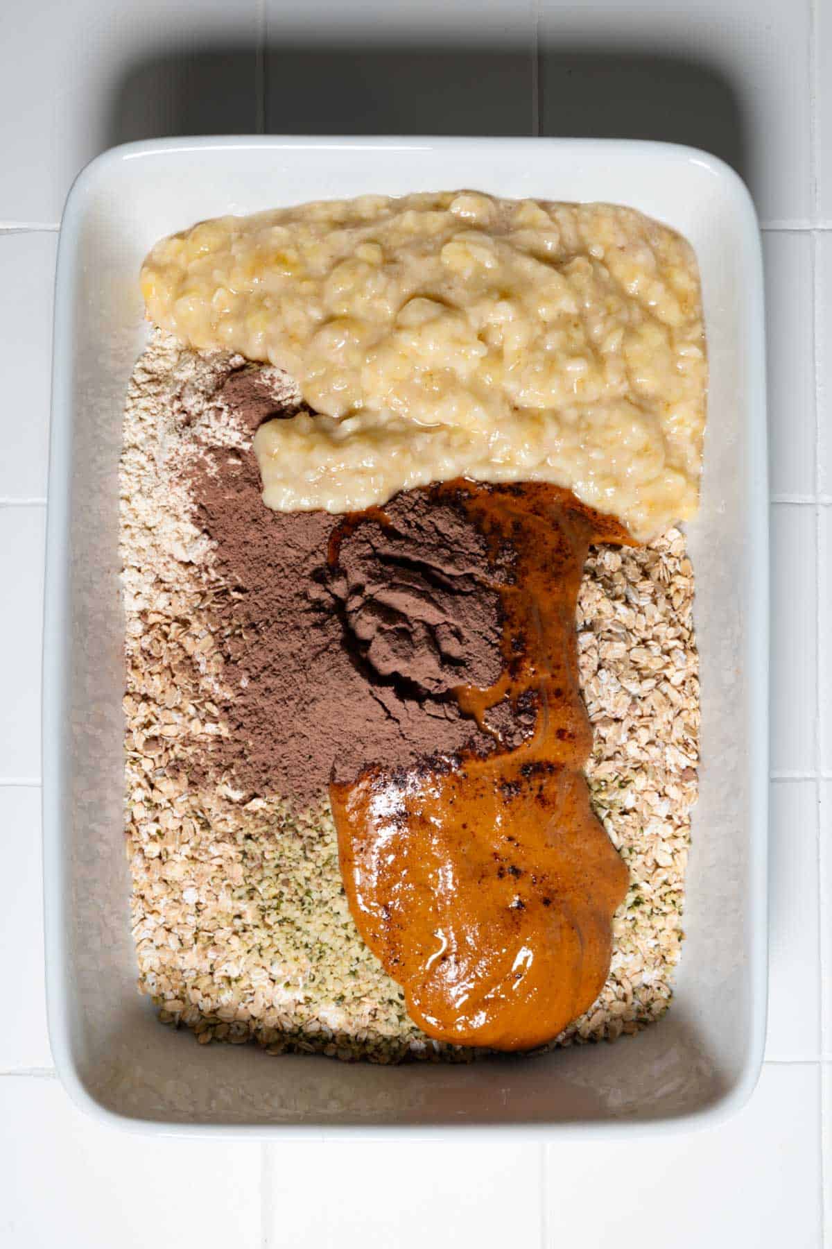 Mashed banana, oats, nut butter, hemp hearts, cocoa powder, and protein powder in a white baking dish.