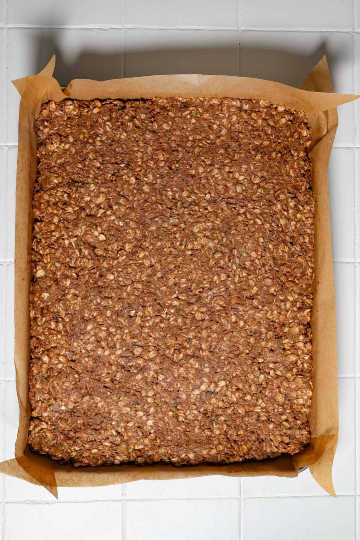Mashed oats, banana, cocoa powder, and hemp hearts pressed into a rectangular baking dish on parchment paper.