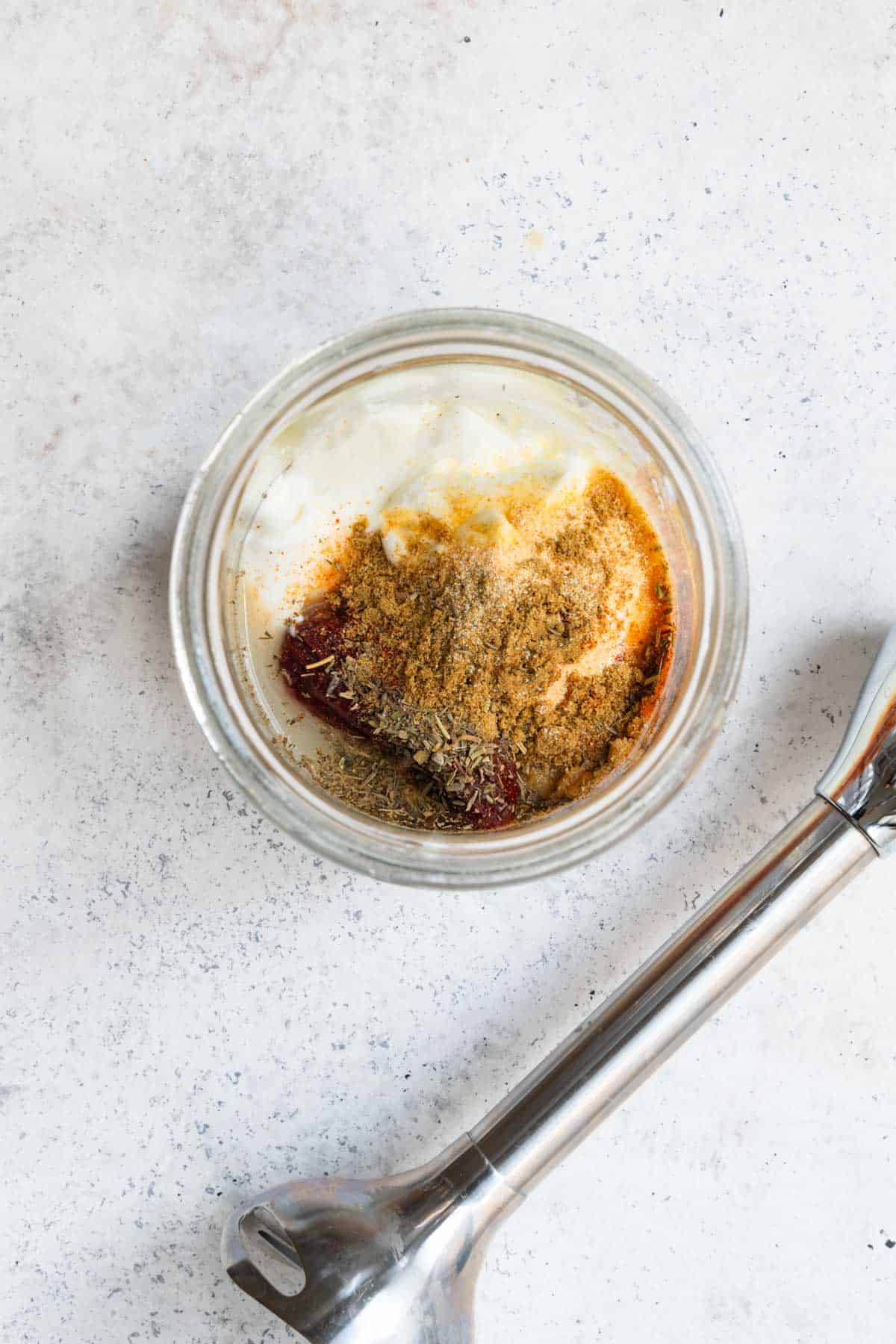 Yogurt, chipotle peppers, seasoning, and lime juice in a glass jar next to an immersion blender.