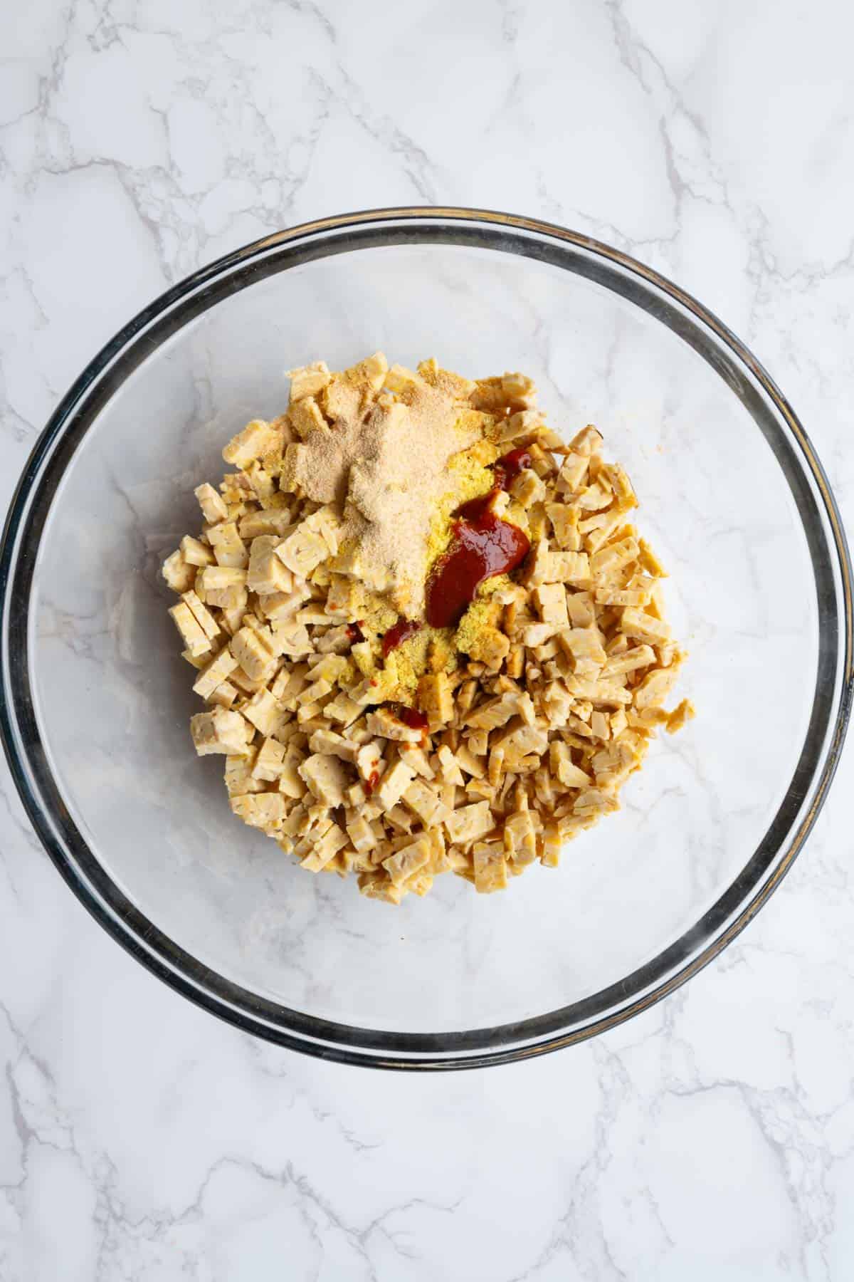 Chopped tempeh, sriracha, nutritional yeast, garlic powder, and tamari in a glass bowl.