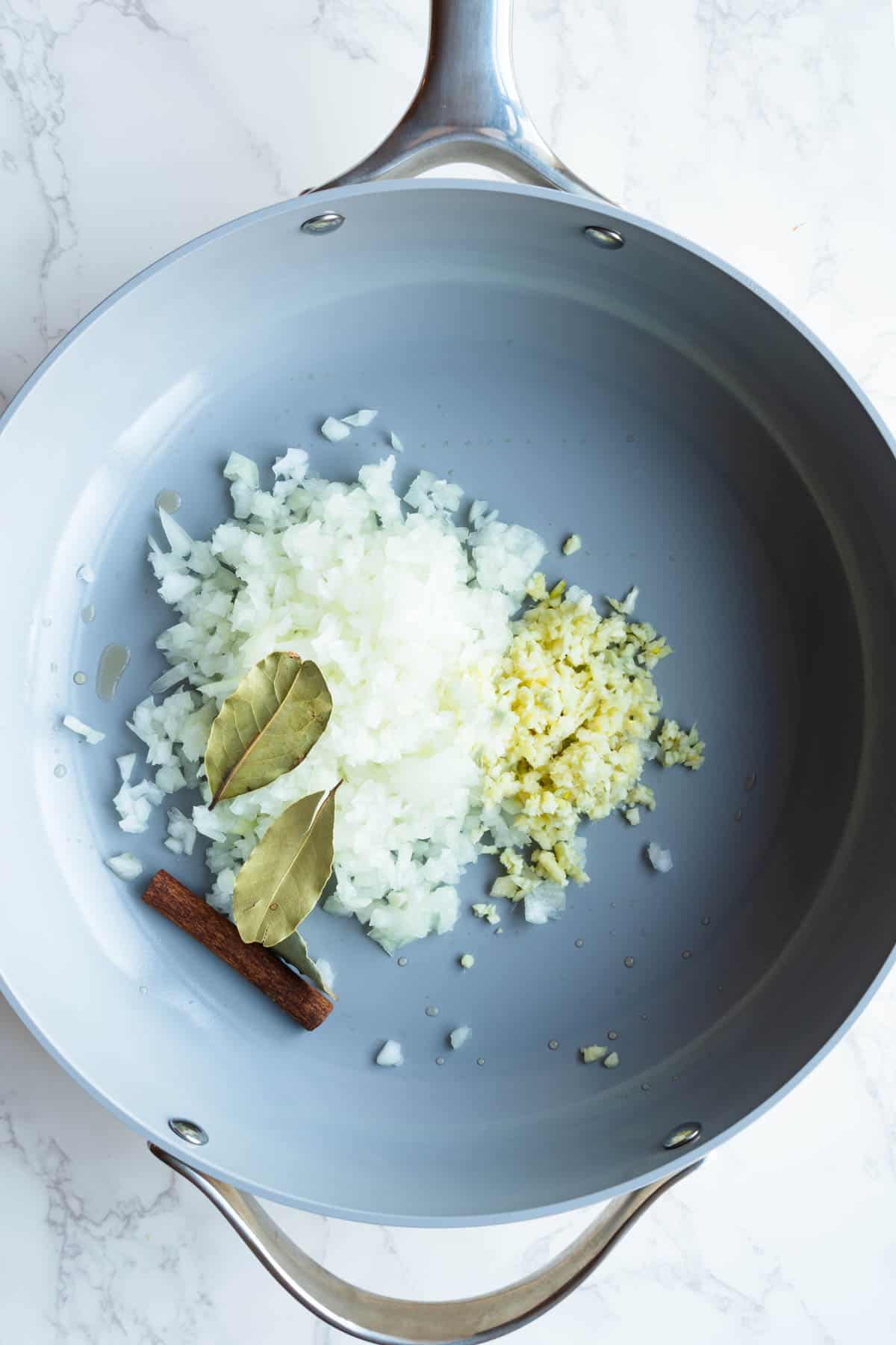 Chopped onion, garlic, ginger, three bay leaves, and a cinnamon stick with a drizzle of broth in a large skillet.
